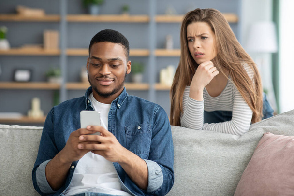 Недоверие. Внимательная леди смотрит на черного парня, пока он пишет смс на смартфоне - Фото, изображение