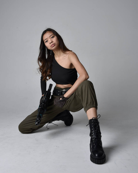 Full length portrait of pretty brunette, asian girl wearing black top and khaki utilitarian army pants and leather boots. Sitting pose holding a science fiction gun, isolated agent a light grey studio background. - Photo, Image