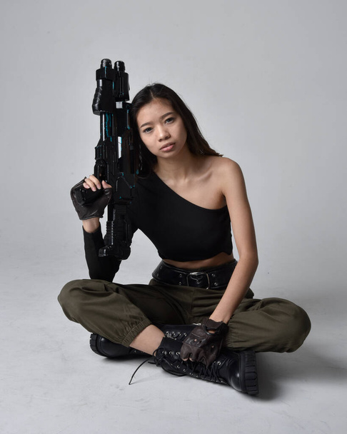 Teljes hosszúságú portré csinos barnáról, ázsiai lányról fekete felsőben és khaki hasznos katonai nadrágban és bőrcsizmában. Ülő póz kezében sci-fi fegyver, elszigetelt ügynök világos szürke stúdió háttér. - Fotó, kép