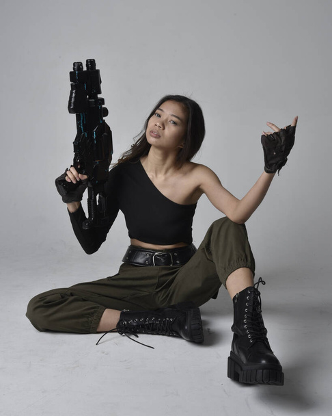 Full length portrait of pretty brunette, asian girl wearing black top and khaki utilitarian army pants and leather boots. Sitting pose holding a science fiction gun, isolated agent a light grey studio background. - Fotografie, Obrázek