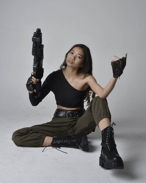 Uzun boylu, esmer, siyah bluz giyen Asyalı bir kız ve asker pantolonu ve deri çizmeleri. Elinde bir bilim kurgu silahı, izole edilmiş bir ajan, açık gri bir stüdyo arka planı var.. - Fotoğraf, Görsel