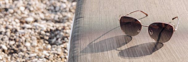 Stylish women's sunglasses with bright sunlight and shadow on background of a pebble beach. Concept of travel, summer vacation and rest on beach. Copy space. Banner - Photo, image