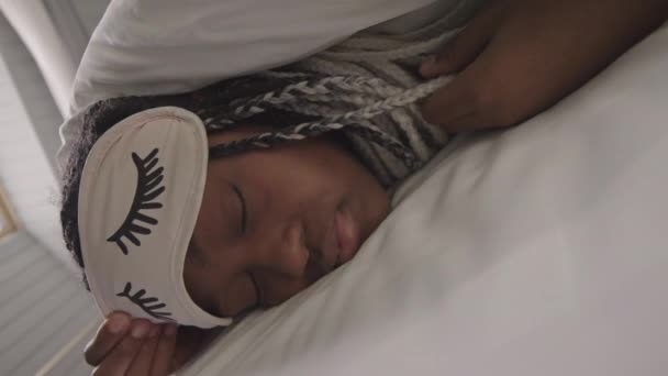 Retrato de cerca de una joven afroamericana mirando a la cámara acostada en la cama con pestañas rosas máscara de sueño - Imágenes, Vídeo