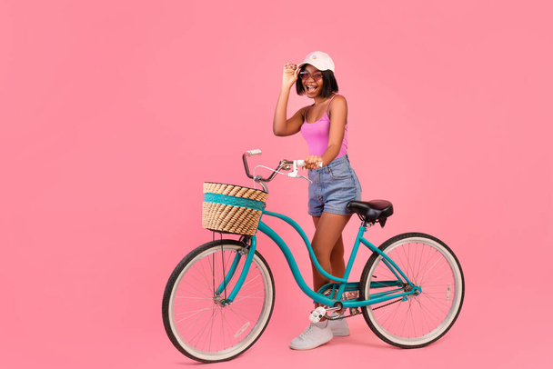 Portrait pleine longueur de dame noire émotionnelle en vêtements d'été debout avec vélo sur fond de studio rose, espace de copie - Photo, image