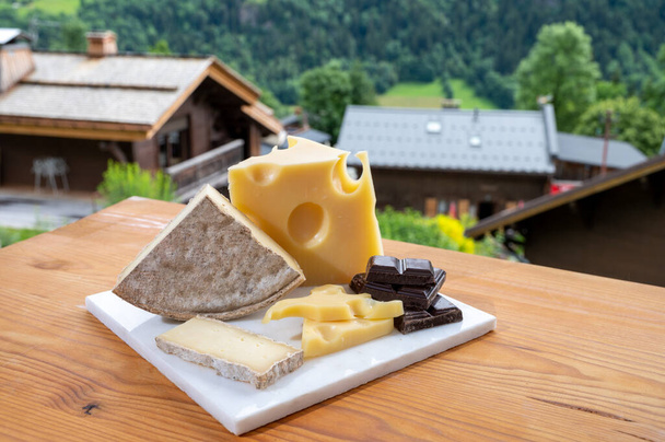 Cheese collection, Swiss cow cheese emmental, tomme and dark chocolate and mountains village in green Apls in summer on background - Photo, Image