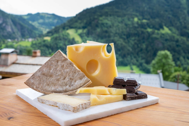 Coleção de queijos, queijo suíço de vaca emmental, tomme e chocolate escuro e montanhas aldeia em verde Apls no verão no fundo - Foto, Imagem