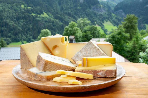 Sbírka sýrů, dřevěná deska s francouzskými sýry comte, beaufort, abondance, emmental, morbier a francouzská horská vesnice v Haute-Savoie v létě na pozadí - Fotografie, Obrázek