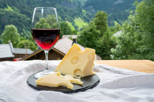 Käsekollektion, Emmentaler französischer Kuhkäse, Glas Rotwein aus Savoyen und französisches Bergdorf in Haute-Savoie im Sommer im Hintergrund - Foto, Bild