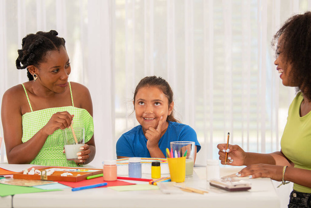 Litlle latina girl making funny face in art class with her two teachers while painting. - Photo, Image