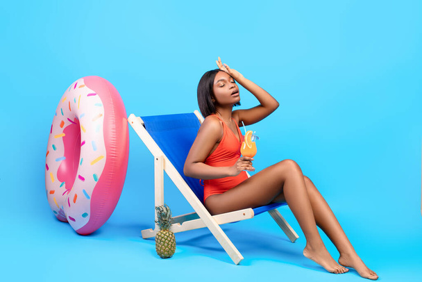 Повна довжина молодої чорної леді, яка страждає від літньої спеки, тримає тропічний коктейль, сидячи в кріслі для відпочинку
 - Фото, зображення