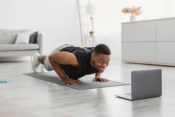 Атлетик Африканский парень упражнения на ноутбуке делает толчки-Ups дома - Фото, изображение
