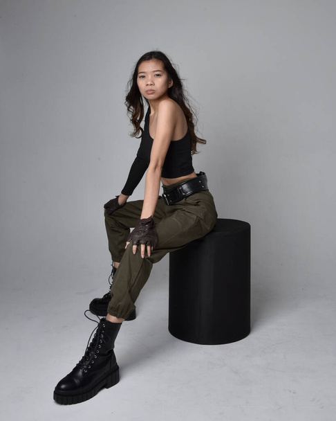 Full length portrait of pretty brunette, asian girl wearing black top and khaki utilitarian army pants and leather boots. Sitting pose , isolated agent a light grey studio background. - Fotó, kép