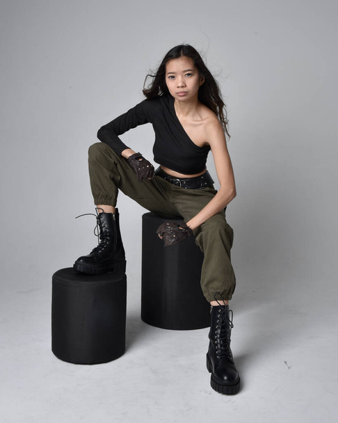 Full length portrait of pretty brunette, asian girl wearing black top and khaki utilitarian army pants and leather boots. Sitting pose , isolated agent a light grey studio background. - Foto, imagen