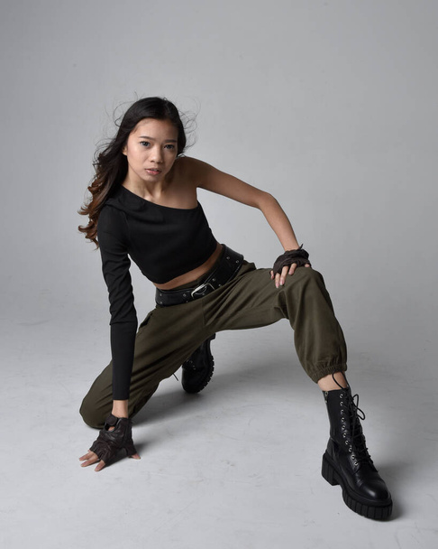 Full length portrait of pretty brunette, asian girl wearing black top and khaki utilitarian army pants and leather boots. Sitting pose , isolated agent a light grey studio background. - Photo, Image