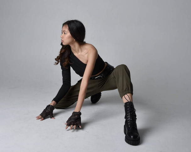 Full length portrait of pretty brunette, asian girl wearing black top and khaki utilitarian army pants and leather boots. Sitting pose , isolated agent a light grey studio background. - Foto, Imagem