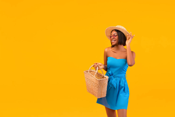 Summer is here. Portrait of young black lady in fashionable outfit going to beach with straw bag on orange background - Foto, Imagen