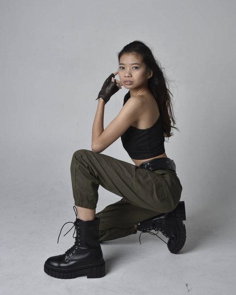 Full length portret van mooie brunette, aziatische meisje dragen zwarte top en kaki utilitaristische leger broek en lederen laarzen. Zittende houding, geïsoleerd agens een lichtgrijze studio achtergrond. - Foto, afbeelding