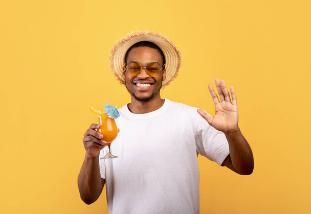 Привлекательный черный парень в футболке и соломенной шляпе держит стакан свежего коктейля и машет перед камерой на желтом фоне - Фото, изображение