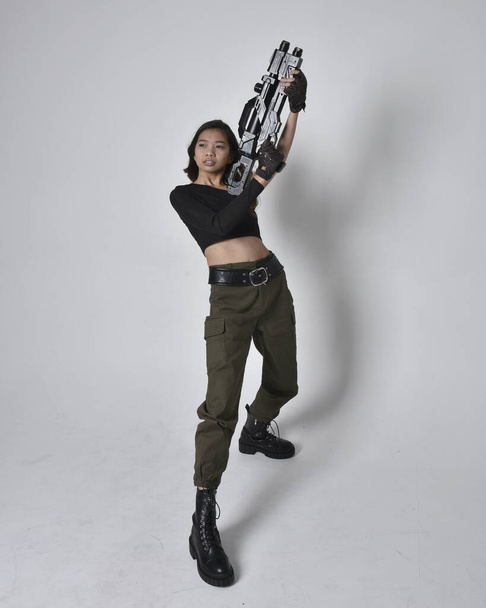Full length portrait of pretty brunette, asian girl wearing black top and khaki utilitarian army pants and leather boots. Standing pose holding a science fiction gun, isolated agent a light grey studio background. - Фото, изображение