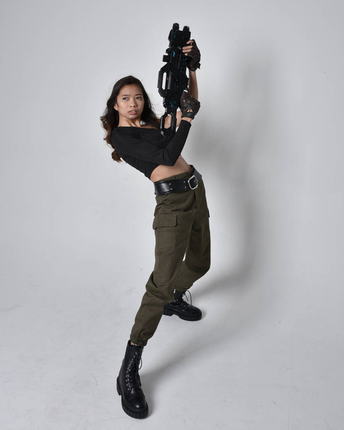 Full length portret van mooie brunette, aziatische meisje dragen zwarte top en kaki utilitaristische leger broek en lederen laarzen. Stand houden van een sciencefiction pistool, geïsoleerde agent een lichtgrijze studio achtergrond. - Foto, afbeelding