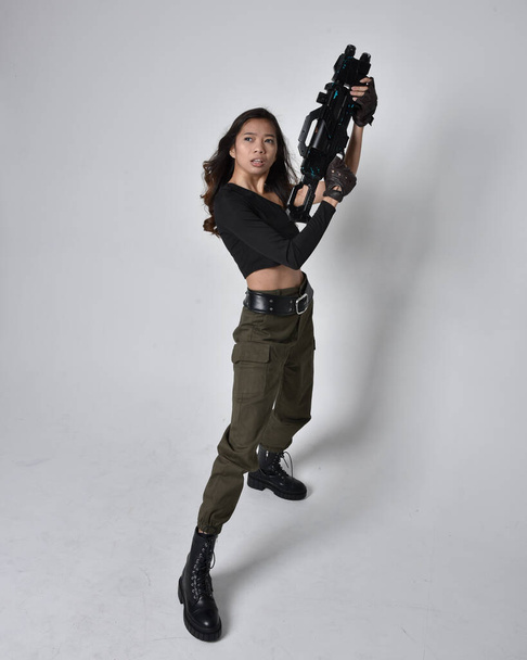 かなりブルネットの完全な長さの肖像画、黒の上とカーキ実用的な軍のズボンと革のブーツを身に着けているアジアの女の子。SF銃を持っている立ちポーズ、孤立したエージェントライトグレーのスタジオの背景. - 写真・画像