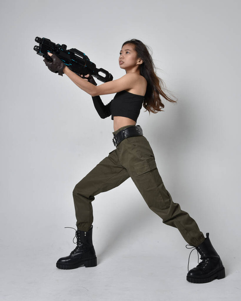 Full length portrait of pretty brunette, asian girl wearing black top and khaki utilitarian army pants and leather boots. Standing pose holding a science fiction gun, isolated agent a light grey studio background. - Valokuva, kuva
