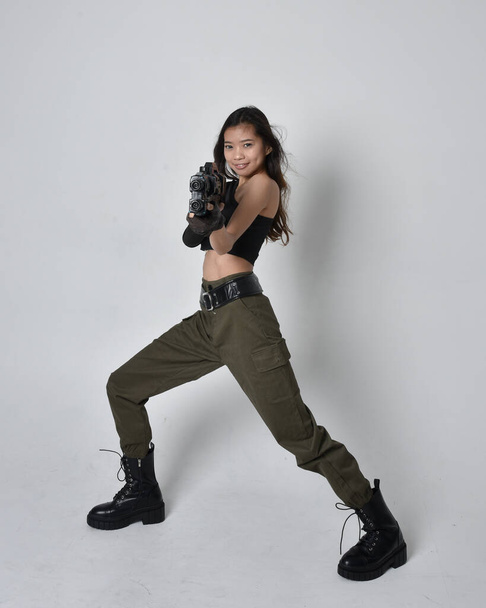 Full length portrait of pretty brunette, asian girl wearing black top and khaki utilitarian army pants and leather boots. Standing pose holding a science fiction gun, isolated agent a light grey studio background. - Photo, Image