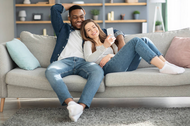 Χαρούμενοι πολυπολιτισμικοί σύζυγοι χαλαρώνουν στον καναπέ με smartphone μαζί - Φωτογραφία, εικόνα