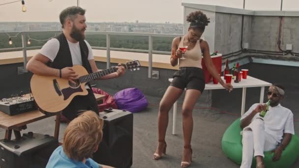 Слоумо отслеживание бородатого молодого человека, играющего на акустической гитаре и поющего песни своим друзьям на террасе на крыше - Кадры, видео