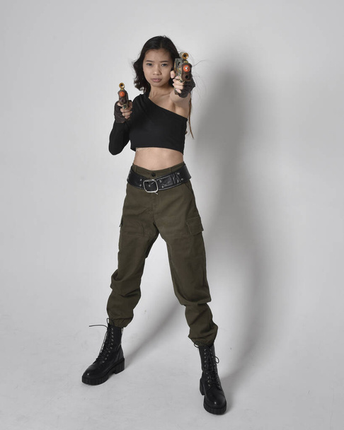 Full length portret van mooie brunette, aziatische meisje dragen zwarte top en kaki utilitaristische leger broek en lederen laarzen. Stand houden van een sciencefiction pistool, geïsoleerde agent een lichtgrijze studio achtergrond. - Foto, afbeelding