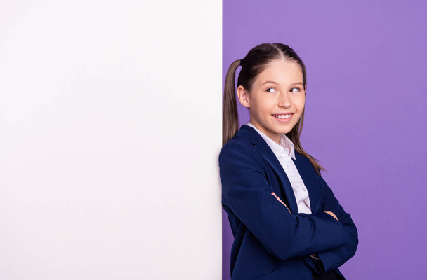Photo of charming funny school girl wear blue uniform arms folded looking white billboard empty space isolated purple color background - Photo, Image