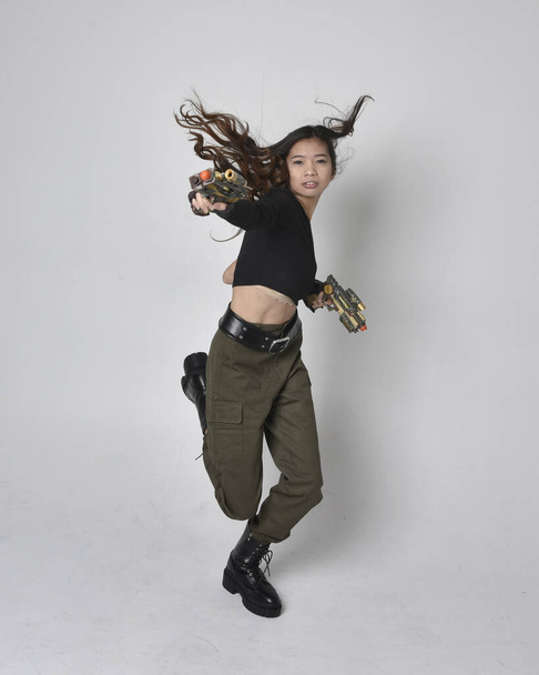 Full length portrait of pretty brunette, asian girl wearing black top and khaki utilitarian army pants and leather boots. Standing pose holding a science fiction gun, isolated agent a light grey studio background. - 写真・画像
