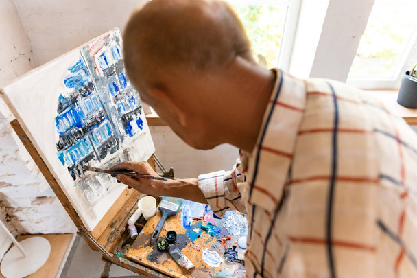 Rijpere of oudere grijze schilder met een borstel voor de camera. Serieuze man in een met verf bevlekt shirt aan het werk in de studio of thuis - Foto, afbeelding