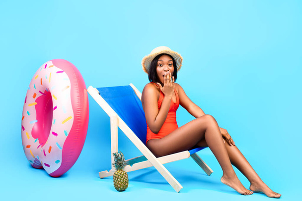 Oops. Shy black lady in bikini chilling in lounge chair, covering her mouth, making mistake on blue studio background - Fotó, kép