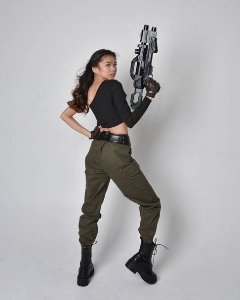 Full length portret van mooie brunette, aziatische meisje dragen zwarte top en kaki utilitaristische leger broek en lederen laarzen. Staande houding met sciencefiction pistool, geïsoleerd tegen studio achtergrond - Foto, afbeelding