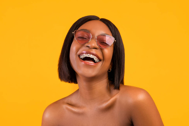 Portrait de femme noire joyeuse en lunettes de soleil riant, exprimant une émotion positive sur fond de studio orange - Photo, image