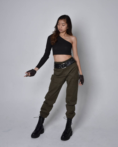 Full length portrait of pretty brunette, asian girl wearing black top and khaki utilitarian army pants and leather boots. Standing pose with gestural hands, isolated against a light grey studio background. - Foto, afbeelding