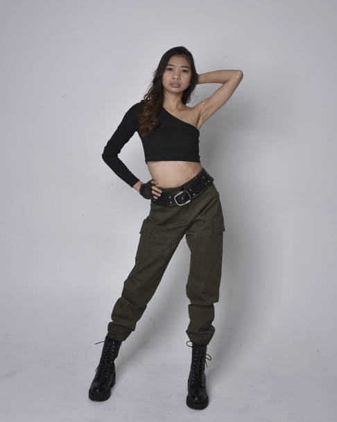 Full length portrait of pretty brunette, asian girl wearing black top and khaki utilitarian army pants and leather boots. Standing pose with gestural hands, isolated against a light grey studio background. - Photo, Image