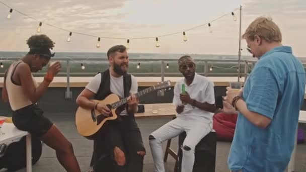 Handheld shot of young man with mobile phone filming during rooftop party with his friends. Bearded man playing acoustic guitar and singing song - Footage, Video