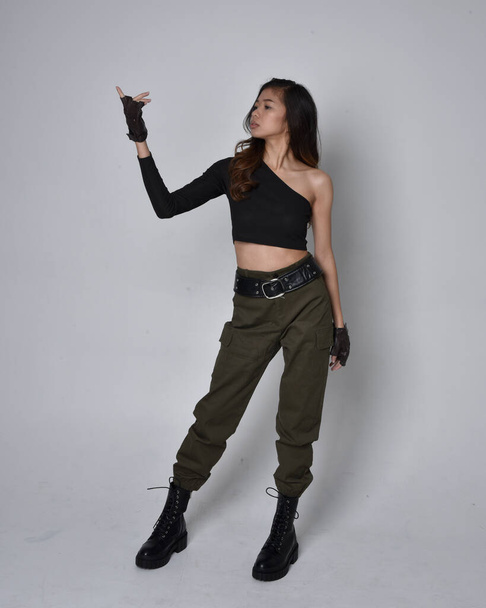 Full length portrait of pretty brunette, asian girl wearing black top and khaki utilitarian army pants and leather boots. Standing pose with gestural hands, walking away from the camera.  isolated against a light grey studio background. - Fotó, kép