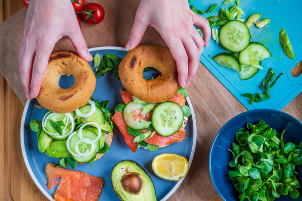 Hands making bagel sandwiches. Gluten free toasted bagels with smoked salmon and cream cheese and with avocado, cucumber, lettuce, and onions. Homemade Healthy breakfast, fun vibrant colors. Top view - Photo, Image