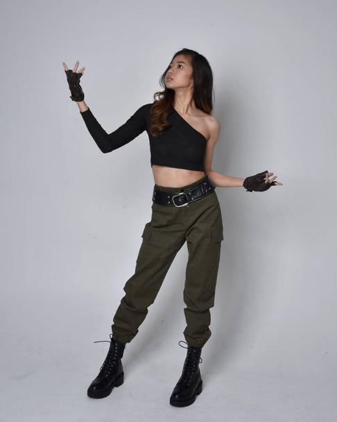 Full length portrait of pretty brunette, asian girl wearing black top and khaki utilitarian army pants and leather boots. Standing pose with gestural hands, walking away from the camera.  isolated against a light grey studio background. - Foto, immagini