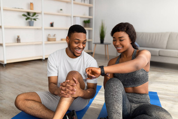 Sportieve zwarte dame toont gegevens op smartwatch of fitness tracker aan haar vriend na thuis workout - Foto, afbeelding