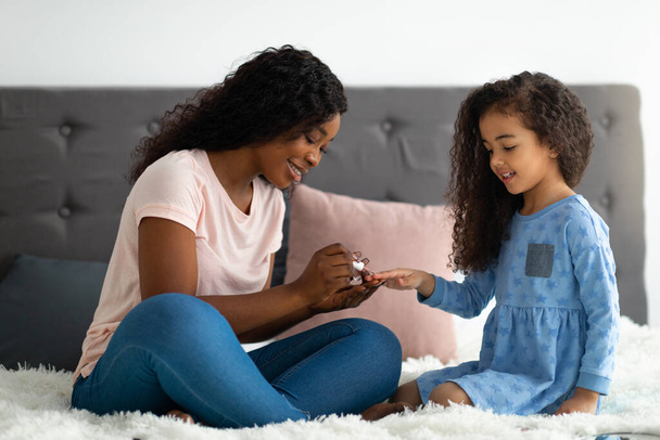 Jeune mère noire appliquant du vernis sur ses adorables filles ongles sur le lit à la maison, pleine longueur - Photo, image