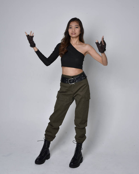 Full length portrait of pretty brunette, asian girl wearing black top and khaki utilitarian army pants and leather boots. Standing pose with gestural hands, walking away from the camera.  isolated against a light grey studio background. - Φωτογραφία, εικόνα