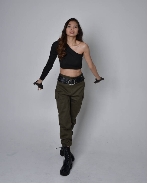 Full length portrait of pretty brunette, asian girl wearing black top and khaki utilitarian army pants and leather boots. Standing pose with gestural hands, walking away from the camera.  isolated against a light grey studio background. - Φωτογραφία, εικόνα
