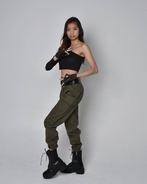 Full length portrait of pretty brunette, asian girl wearing black top and khaki utilitarian army pants and leather boots. Standing pose with gestural hands, walking away from the camera.  isolated against a light grey studio background. - Photo, Image