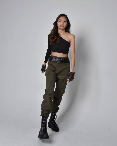 Full length portrait of pretty brunette, asian girl wearing black top and khaki utilitarian army pants and leather boots. Standing pose with gestural hands, walking away from the camera.  isolated against a light grey studio background. - Photo, image