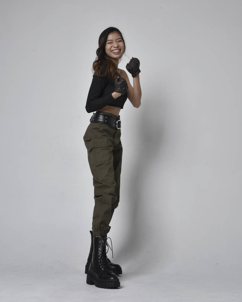 Full length portrait of pretty brunette, asian girl wearing black top and khaki utilitarian army pants and leather boots. Standing pose with gestural hands, walking away from the camera.  isolated against a light grey studio background. - Photo, Image
