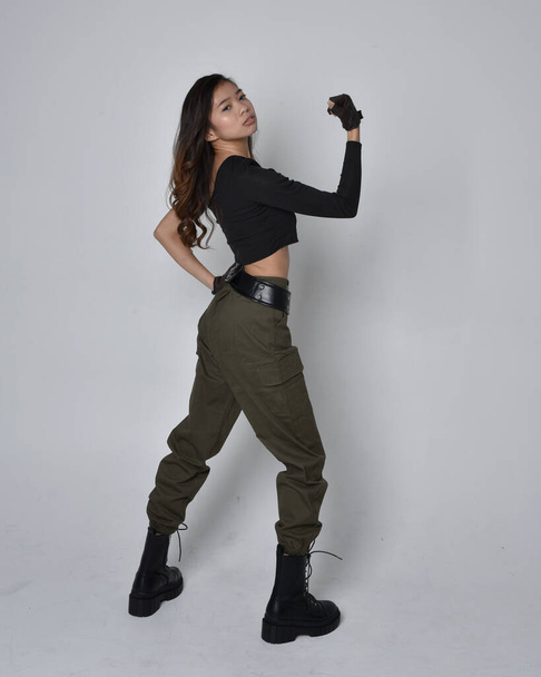 Full length portrait of pretty brunette, asian girl wearing black top and khaki utilitarian army pants and leather boots. Standing pose with gestural hands, walking away from the camera.  isolated against a light grey studio background. - Valokuva, kuva